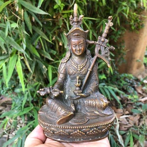 Puede incluir: Estatua de bronce de una figura sentada con corona y ropa ornamentada, sosteniendo un báculo y un pequeño recipiente. La estatua tiene detalles intrincados y está sobre un fondo de follaje verde.