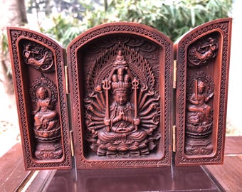 May include: A wooden, three-panel folding screen with detailed carvings. The central panel shows a multi-armed figure in a lotus position, with flames around it. The side panels depict seated figures and celestial beings. The screen is a rich, reddish-brown colour.