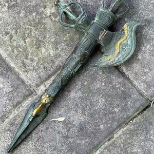 Könnte beinhalten: Eine rituelle Axt aus dunkelgrünem und bronzefarbenem Metall mit einem verzierten Griff, der aufwendige Reliefschnitzereien aufweist. Sie besitzt eine spitze Klinge und ein gebogenes Blatt, das mit einem goldfarbenen Drachenmotiv verziert ist.