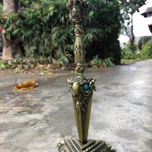 May include: An antique-style dagger with a gold finish. The dagger features a detailed handle with a face design and turquoise accents. It stands on a decorative square base, showcasing intricate patterns. The design suggests a ceremonial or decorative purpose.