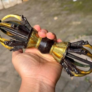 May include: A ritual vajra object featuring a central dark brown handle with a spherical grip, flanked by two symmetrical ends. Each end consists of four curved, golden-yellow prongs that meet at a pointed tip, accented with dark brown metal details.