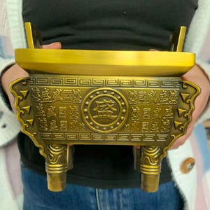 Puede incluir: Un quemador de incienso dorado con intrincadas tallas y caracteres chinos. El quemador tiene forma rectangular con un asa a cada lado y cuatro patas. El diseño central presenta un emblema circular con más caracteres.