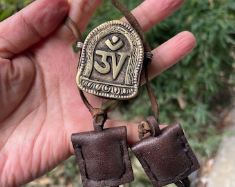 May include: A brass pendant featuring the Om symbol in a raised relief design, attached to a brown leather cord. Two small, square, dark brown leather pouches are secured to the cord below the pendant with visible black stitching.