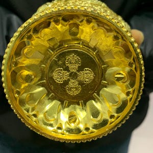 May include: A gold-colored, ornate vessel with intricate patterns and designs. The base features a detailed emblem, and the overall aesthetic suggests a ceremonial or decorative purpose. The vessel is held from the bottom, showcasing its detailed craftsmanship.