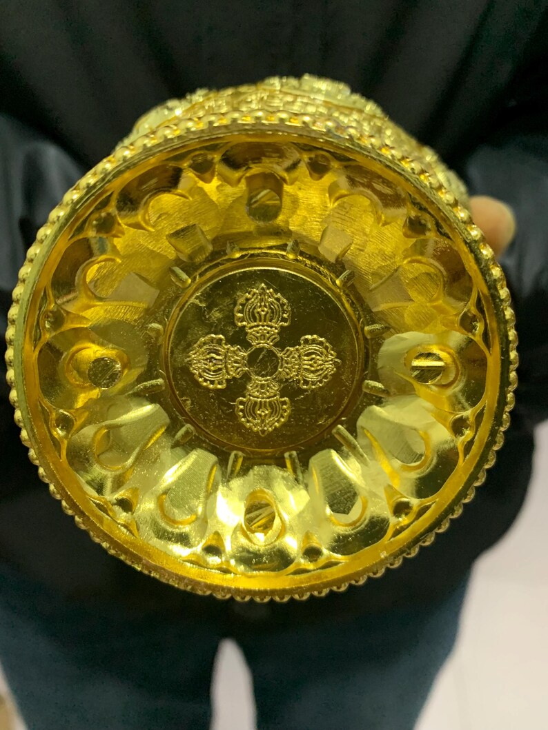 May include: A gold-colored, ornate vessel with intricate patterns and designs. The base features a detailed emblem, and the overall aesthetic suggests a ceremonial or decorative purpose. The vessel is held from the bottom, showcasing its detailed craftsmanship.