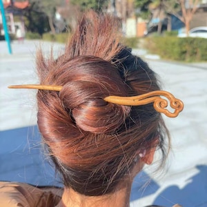 May include: A wooden hair stick with a cloud-shaped design. The hair accessory is inserted into a bun of dark brown hair. The hair stick is a light brown color and appears to be made of wood.
