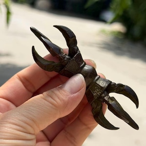 May include: A dark bronze-coloured metal vajra ritual object featuring a central handle and two symmetrical ends, each composed of three curved, pointed prongs that meet at a central tip, displaying intricate engraved patterns on the base.