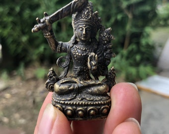 May include: A small, bronze-coloured statue of a seated deity, holding a sword aloft in one hand and displaying a mudra with the other. The figure is adorned with intricate details, including a crown and ornate jewellery. The statue is likely a representation of a religious figure.