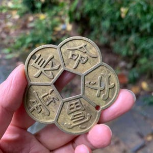 May include: A vintage brass coin with a unique flower-like shape. The coin features five sections, each inscribed with an Asian character. The coin has a central hole and a small hole at the edge. The coin is held in a hand against a blurred green background.