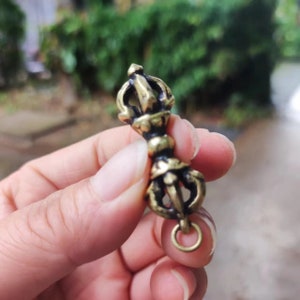 May include: An antique-style brass vajra, a ritual object with intricate, symmetrical designs. The vajra has a central handle with decorative elements and a ring at the base. The object is held in a hand against a blurred green backdrop.
