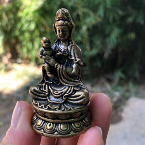 Può includere: Una statua color bronzo di una figura seduta che tiene in braccio un bambino. La statua presenta dettagli intricati, tra cui gli abiti e i capelli della figura, e la base su cui è seduta. Lo sfondo è sfocato, con fogliame verde.
