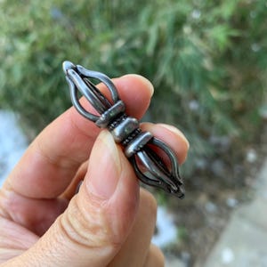 May include: A silver-coloured metal vajra, a ritual object, is held in a hand. The vajra has a central cylindrical section with decorative bands and flared prongs extending from each end. The background is blurred green foliage.