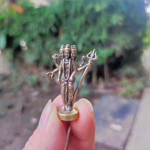 May include: A small, bronze-colored statue of a deity with multiple heads and arms, holding various symbolic objects. The statue is standing on a circular base and has intricate details. The background is blurred, with green foliage.