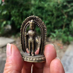 Può includere: Piccola statua di Buddha color bronzo con dettagli intricati. La figura è all'interno di un arco ornato, tenuta in una mano su uno sfondo verde sfocato. La statua presenta una corona e vesti fluenti, mostrando un'espressione serena.