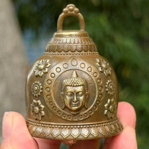 Op de afbeelding: Een messingkleurige metalen bel met een centraal reliëf van een Boeddha-hoofd omringd door een cirkelvormige rand van kleine stippen. De bel is versierd met meerdere bloemmotieven en heeft een geschulpt patroon aan de boven- en onderrand.