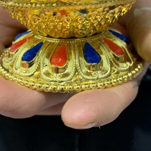 May include: Close-up of a gold-colored decorative bowl with intricate detailing. The base features alternating red and blue teardrop-shaped accents. The bowl has a textured, ornate design, with a shiny, reflective surface.