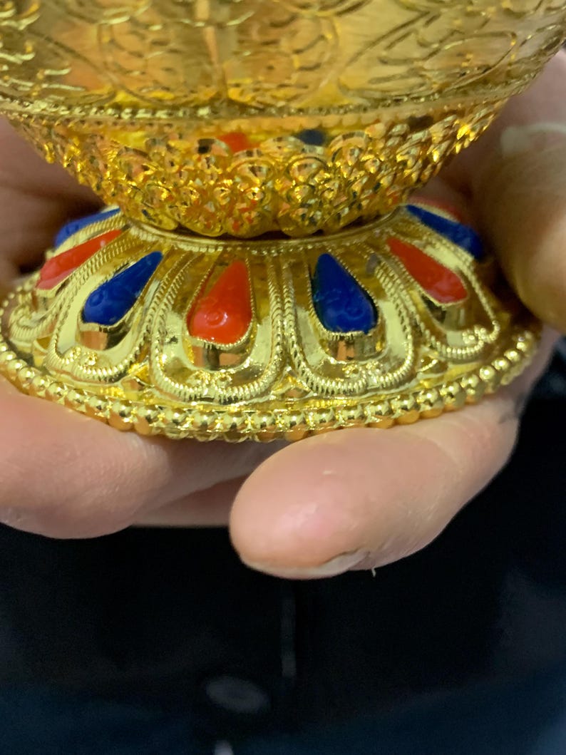 May include: Close-up of a gold-colored decorative bowl with intricate detailing. The base features alternating red and blue teardrop-shaped accents. The bowl has a textured, ornate design, with a shiny, reflective surface.