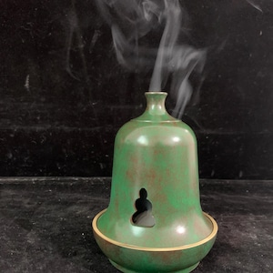 Puede incluir: Quemador de incienso verde y bronce, con forma de campana. Tiene una pequeña abertura frontal y un borde dorado. Humo blanco se eleva desde la parte superior, indicando su uso. El fondo es oscuro. El diseño recuerda a una campana tradicional.