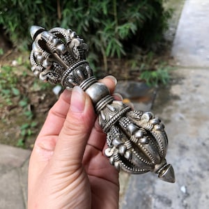 Peut inclure: Objet rituel argenté orné de sculptures détaillées. Il présente une poignée centrale cylindrique, des motifs complexes et se termine par une pointe conique et un motif en forme de couronne. L'objet suggère un usage cérémoniel ou décoratif.