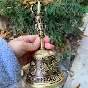 May include: A bronze-colored bell with intricate carvings and a detailed handle featuring a small figure. The bell has a flared base with embossed designs and a decorative top. The overall design suggests a religious or ceremonial item.