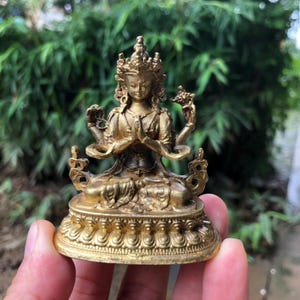 Può includere: Piccola statua dorata di una divinità seduta con più braccia e una corona. La figura è in posa meditativa, con le mani giunte in preghiera. La statua è finemente dettagliata e poggia su una base decorativa. Lo sfondo è un fogliame verde sfocato.