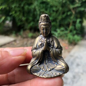 Può includere: Piccola statua color bronzo di una figura seduta con le mani giunte in preghiera. La figura indossa un copricapo dettagliato e vesti fluenti. La statua è alta circa 5 cm, con dettagli intricati.