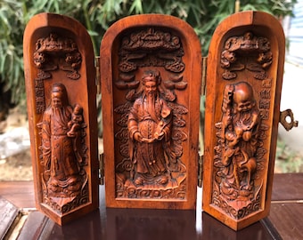 May include: A wooden triptych with carved Chinese deities. The reddish-brown wooden panels are hinged and display detailed relief carvings of robed figures with elaborate facial features and beards. The central panel is slightly larger than the side panels.