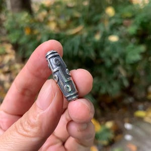 May include: A silver-colored, cylindrical bead with a detailed pattern of circles and lines. The bead is held in a hand against a blurred green and brown background. The bead's design features a series of circular and linear elements.