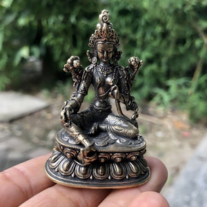 May include: A bronze-coloured statue of a seated deity, with detailed ornamentation. The figure wears a crown and jewellery, and sits on a lotus flower base. The statue is held in a hand, with a blurred green background.