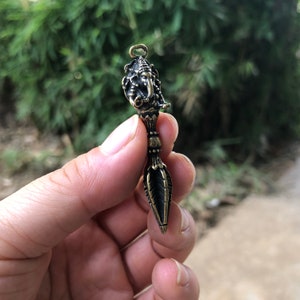 May include: A bronze pendant depicting the head of Ganesha, the Hindu deity, on a decorative handle. The pendant has detailed carvings and a small loop. The background is blurred green foliage.