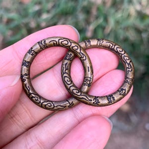 May include: Two bronze rings with detailed cloud and script patterns. The circular rings are made of metal and have an aged appearance. The intricate carvings suggest a vintage or antique style, with each ring's diameter approximately 3 cm.