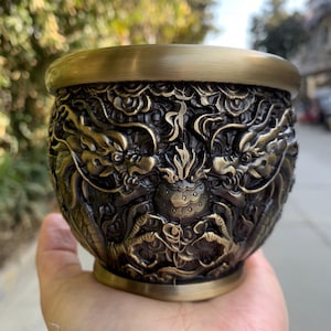 Könnte beinhalten: Bronze-farbene dekorative Schale mit aufwendigen Drachen-Schnitzereien. Die Schale hat eine runde Form mit einem breiten, flachen Rand. Die detaillierte Reliefarbeit zeigt zwei Drachen, die sich gegenüberstehen, mit einem zentralen Gestaltungselement. Die Schale wird in einer Hand gehalten.