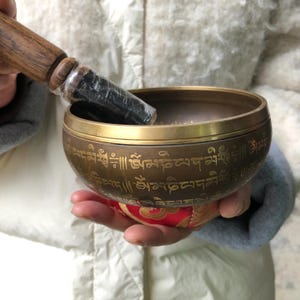 May include: A bronze singing bowl with gold accents and etched script. The bowl is held in a person's hands, resting on a red and gold cushion. A wooden mallet is included, wrapped in plastic.