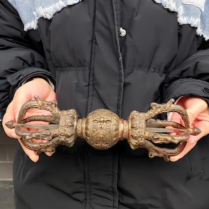 Könnte beinhalten: Ein bronzefarbener Vajra, ein rituelles Objekt, wird in Händen gehalten. Der Vajra weist verzierte, drachenartige Designs an jedem Ende und eine zentrale Kugel mit detaillierten Schnitzereien auf. Das Design deutet auf ein religiöses oder zeremonielles Artefakt hin.