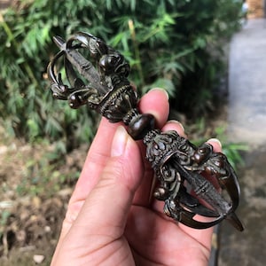 Può includere: Un Vajra color bronzo, un oggetto rituale con dettagli ornati. L'impugnatura presenta una forma bulbosa e le estremità sono progettate con motivi intricati. L'oggetto è tenuto contro uno sfondo verde sfocato.