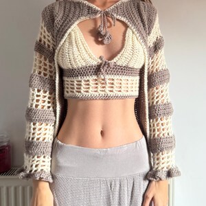 May include: A cream and taupe crochet top and shrug set. The top features a halter neck design with a keyhole detail and small pom-poms. The shrug has long sleeves with a lattice pattern and ruffled cuffs. The set is handmade and suitable for layering.