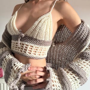 May include: A handmade crochet top and cardigan set in cream and gray. The top features a triangle bralette design with thin straps and a decorative closure. The cardigan has long sleeves with a flared, openwork pattern, creating a bohemian aesthetic.