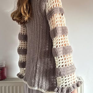 May include: A handmade crochet sweater in shades of gray and cream. The sweater features a solid gray body, cream-colored openwork sleeves with gray accents, and a ruffled hem. The sleeves have a bell-like shape.