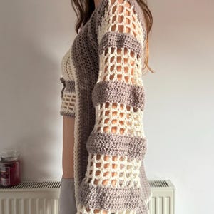 May include: Crocheted top in cream and taupe. The open-weave sleeves have horizontal bands of taupe and cream. The top has a cropped design with a scalloped hem. The garment is handmade and has a bohemian aesthetic.