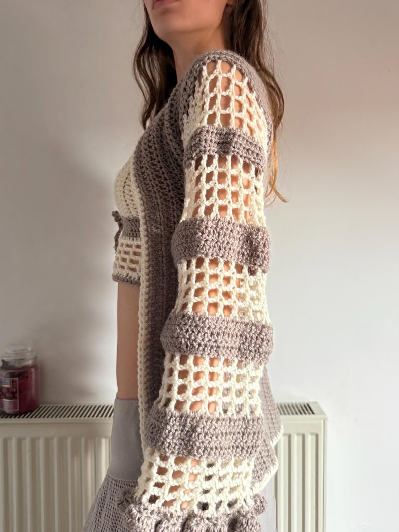 May include: Crocheted top in cream and taupe. The open-weave sleeves have horizontal bands of taupe and cream. The top has a cropped design with a scalloped hem. The garment is handmade and has a bohemian aesthetic.