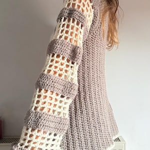 May include: A crocheted sweater in shades of beige and gray. The sleeves feature a unique openwork design with alternating sections of solid and open crochet. The cuffs have a ruffled edge. The body of the sweater has a ribbed texture.