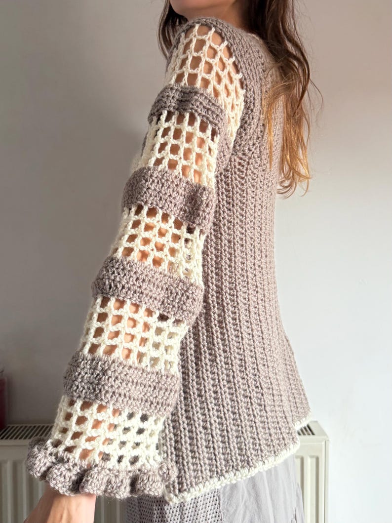 May include: A crocheted sweater in shades of beige and gray. The sleeves feature a unique openwork design with alternating sections of solid and open crochet. The cuffs have a ruffled edge. The body of the sweater has a ribbed texture.