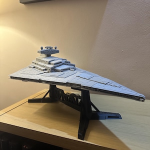 May include: A grey Star Destroyer model, with detailed features, is presented on a black stand. The model is a replica of a spaceship. The model is displayed on a wooden surface. The model is a collectible item.