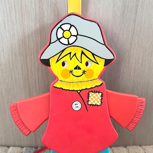 Puede incluir: Un juguete de espantapájaros de plástico vintage y colorido con una cara amarilla, pelo de paja y un sombrero azul. El espantapájaros lleva una camisa roja y pantalones azules. Tiene un lazo amarillo en la parte superior para colgar.