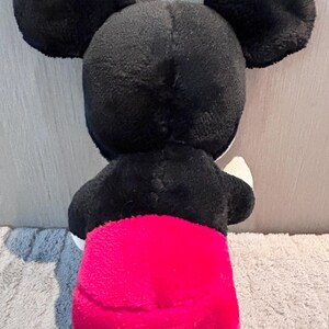 May include: A black and red plush Mickey Mouse toy. The toy is standing on a light brown surface.