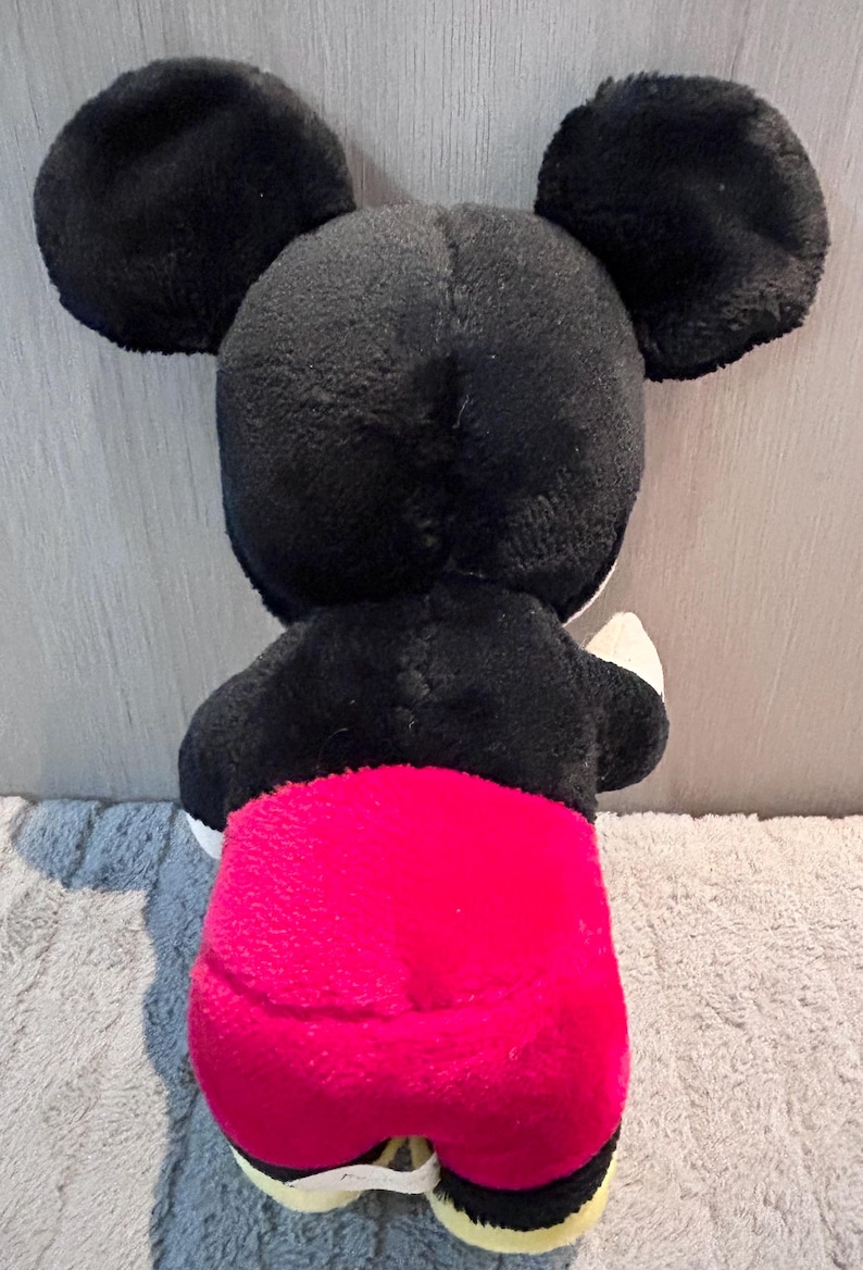 May include: A black and red plush Mickey Mouse toy. The toy is standing on a light brown surface.