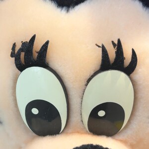 May include: Close-up of a plush toy's face. The toy has large, black eyes with white pupils and black eyelashes. The face is pink and fuzzy.