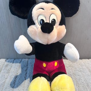 May include: A plush Mickey Mouse toy with black ears, a black nose, and yellow shoes. He is wearing red shorts with two yellow buttons.