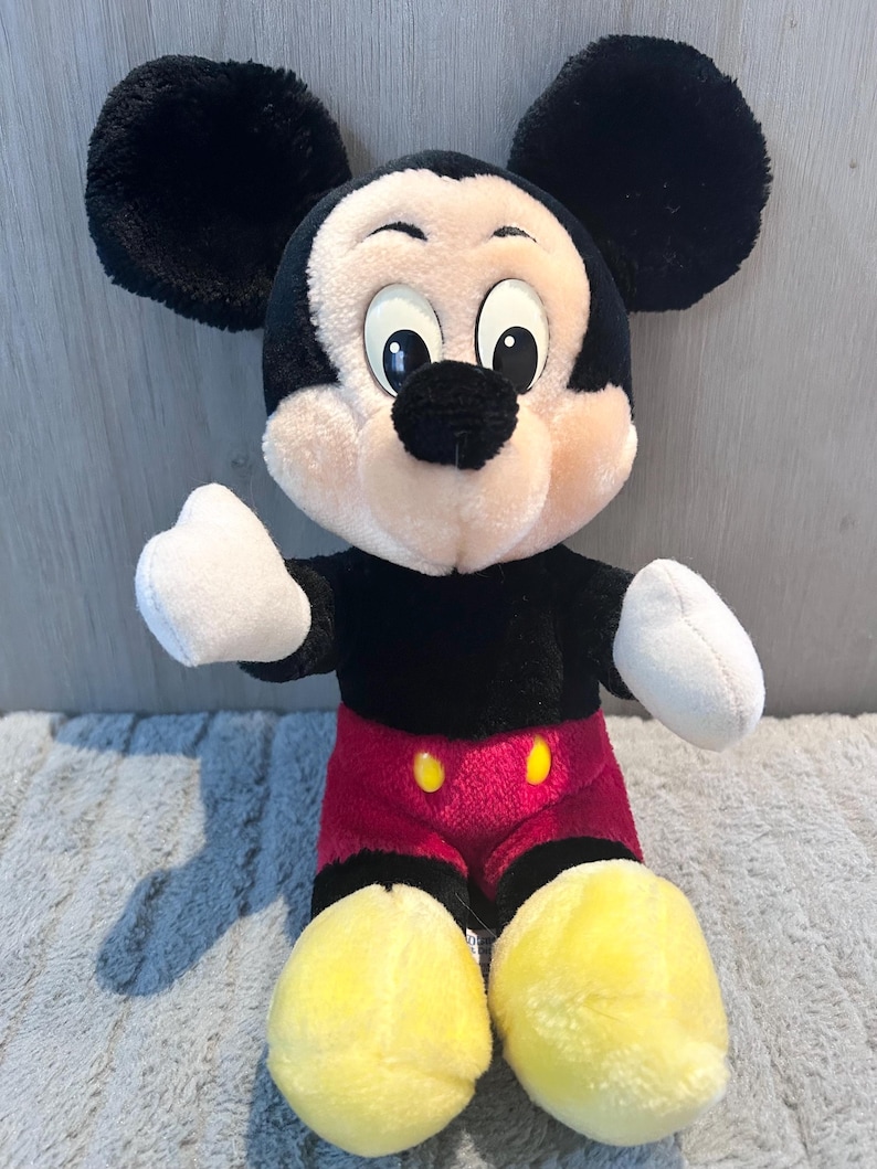 May include: A plush Mickey Mouse toy with black ears, a black nose, and yellow shoes. He is wearing red shorts with two yellow buttons.