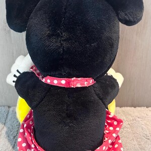 May include: A black and white plush toy of Minnie Mouse wearing a red dress with white polka dots and a red bow with white polka dots.